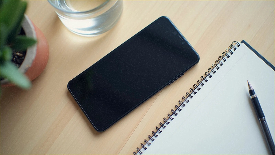 机の上に置かれた電源オフのスマホと、開いたノートとペンです。